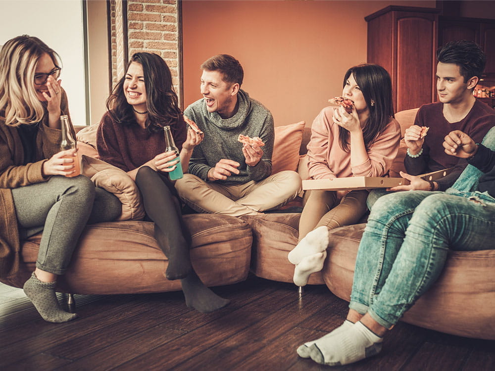 Gruppe von Menschen sitzt bei einer Hausparty auf einem Sofa, trinkt Alkohol und isst Pizza
